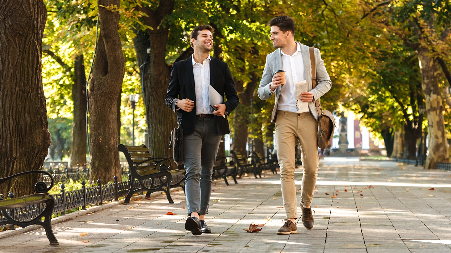 Two men walking down a park path with sun rays breaking through the trees.