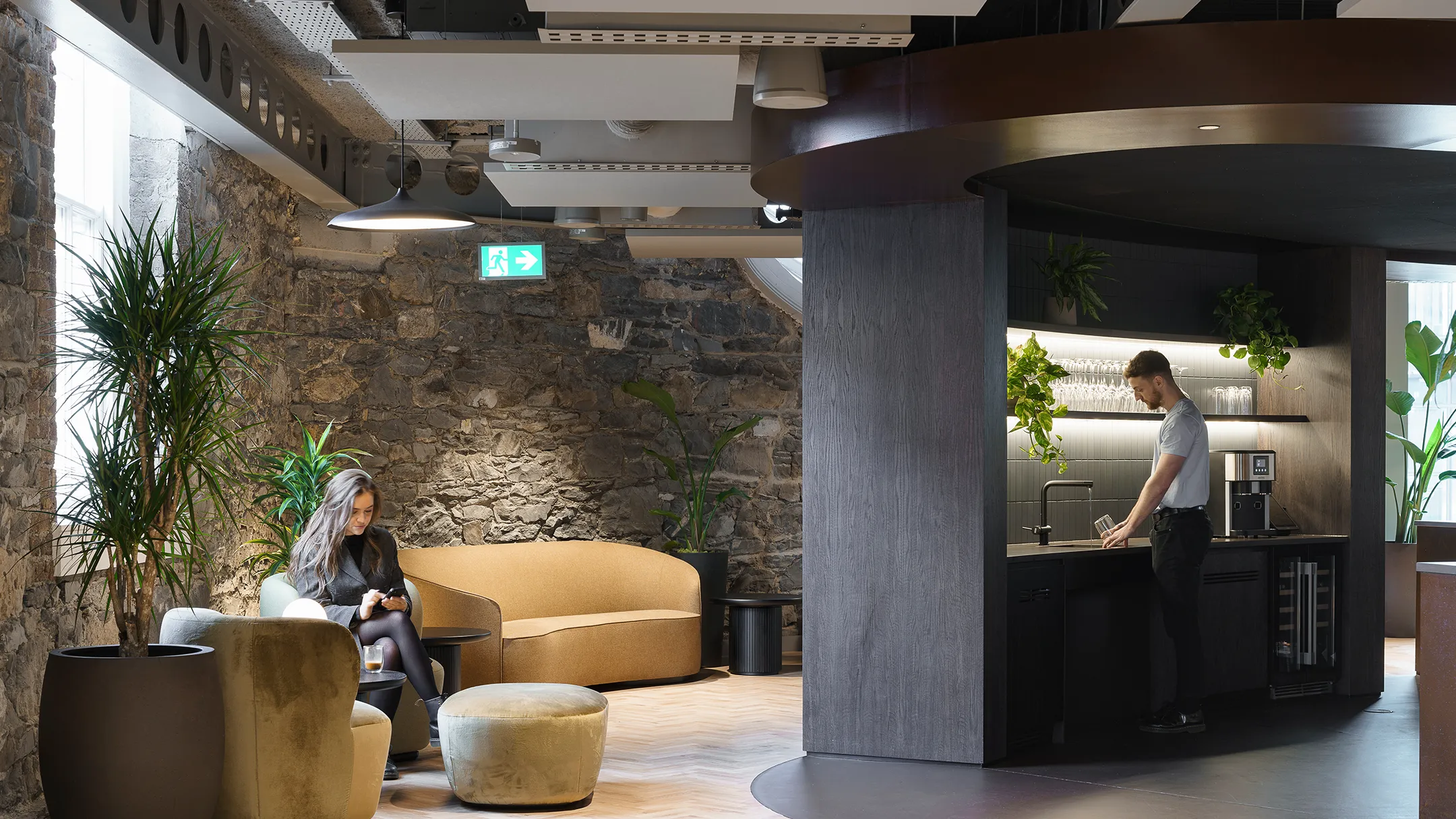 Woman on her phone with coffee sat near a workspace kitchen with a man filling a glass of water.