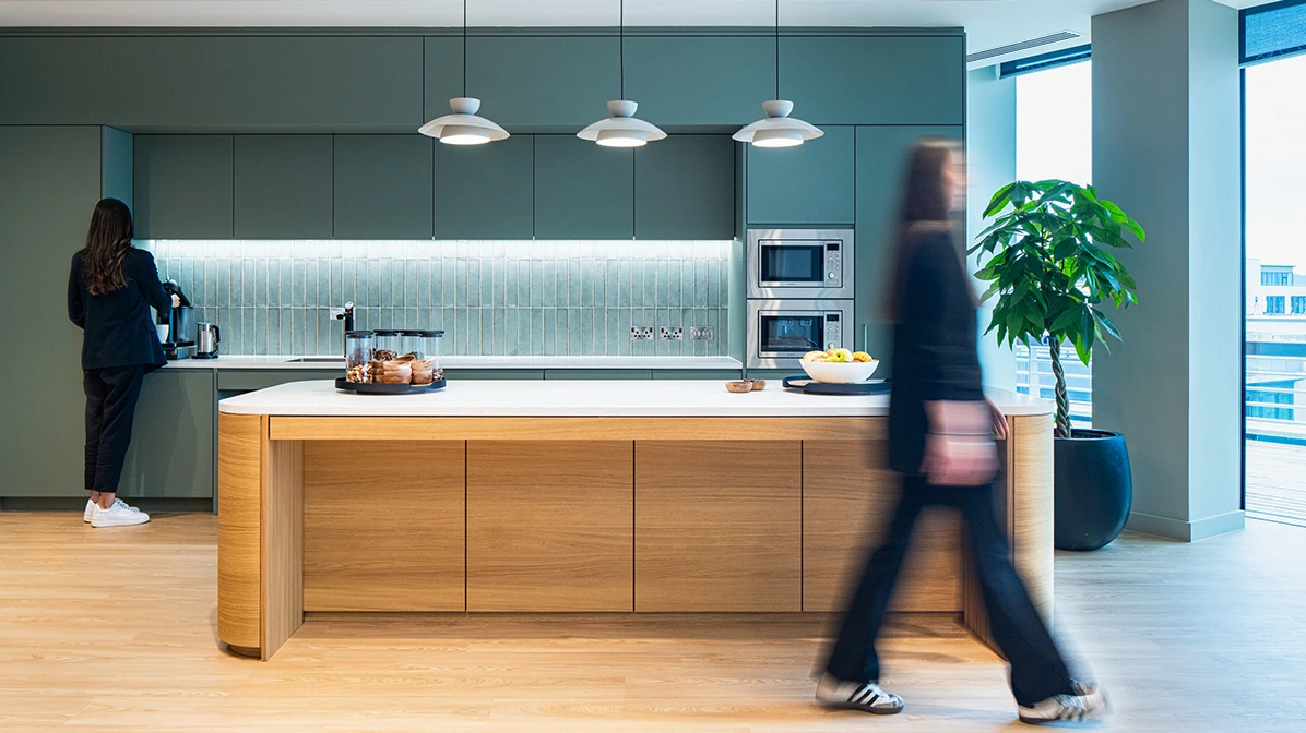 Workspace kitchen with one woman getting a coffee with a second woman blurred in the foreground walking past.